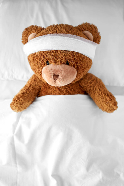 ill teddy bear toy with bandaged head lying in bed - Fotoğraf, Görsel