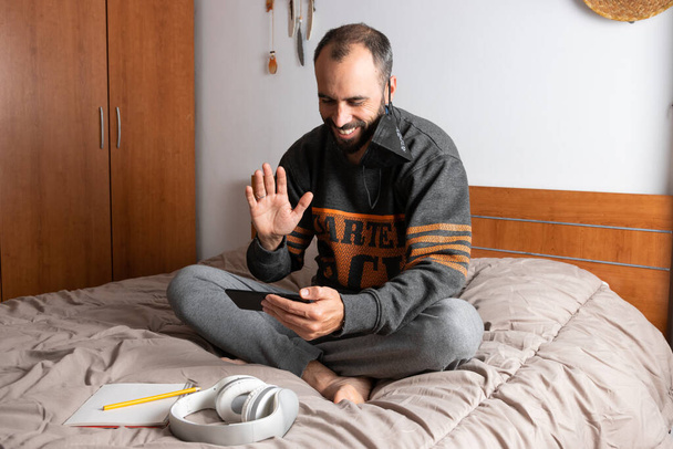 Hombre con auriculares, teléfono móvil y mascarilla, sentado en su cama y confinado a su habitación - Foto, Imagen