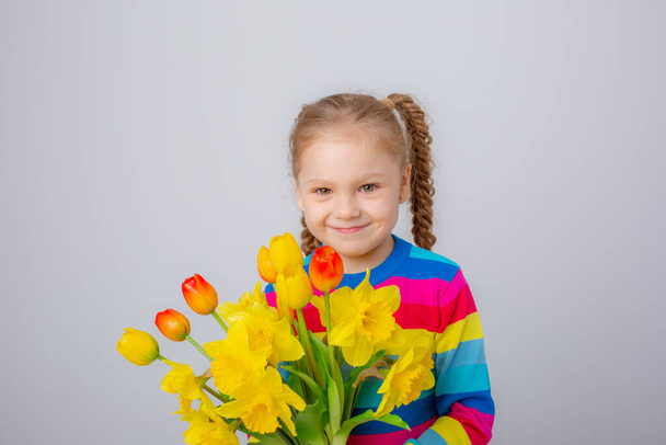 una linda niña en un suéter multicolor sostiene un ramo de flores de primavera sobre un fondo blanco - Foto, Imagen