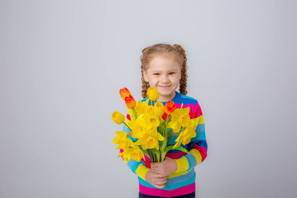 a cute little girl in a multicolored sweater holds a bouquet of spring flowers on a white background - Foto, afbeelding