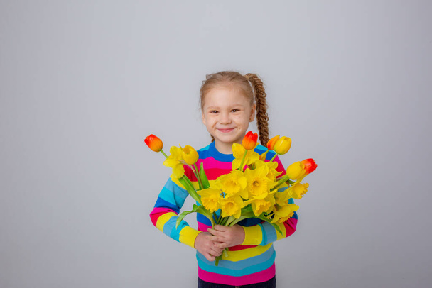 a cute little girl in a multicolored sweater holds a bouquet of spring flowers on a white background - Foto, afbeelding