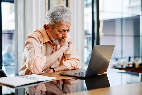 Uno studente anziano è seduto in mensa e segue le lezioni online con attenzione sul suo computer portatile. - Foto, immagini