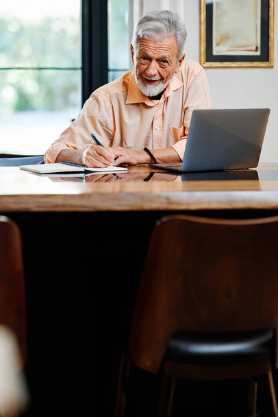 Um estudante sênior feliz está sentado em casa e escrevendo notas em um caderno enquanto segue uma palestra on-line sobre o laptop. - Foto, Imagem