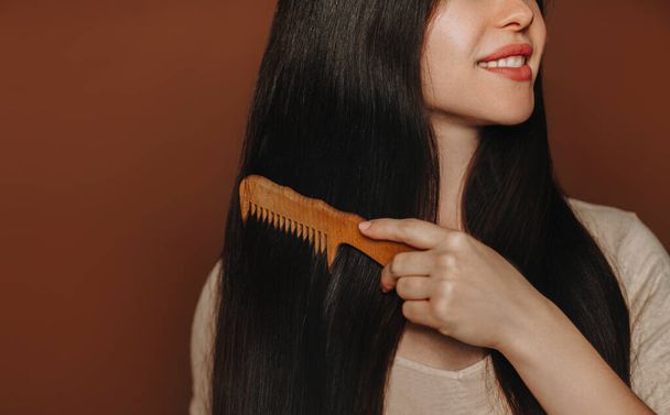 Young happy brunette woman combing her long healthy brown hair and smiling at camera while posing isolated on dark background with copy space for text. Beauty and hair care concept - Foto, Imagen
