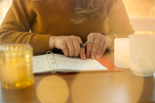 Middle-aged woman writer or poet writing in notebook while sitting at table in sunny room at home, creating novel or poetry. Focused mature female taking notes in planner, planning daily routine - Photo, Image