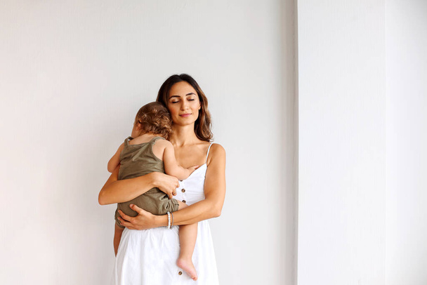 Retrato de maternidad. Joven hermosa madre cariñosa con vestido blanco sosteniendo lindo bebé mientras está de pie aislado sobre la pared blanca. Mamá abrazando pequeño hijo y expresando amor incondicional - Foto, imagen