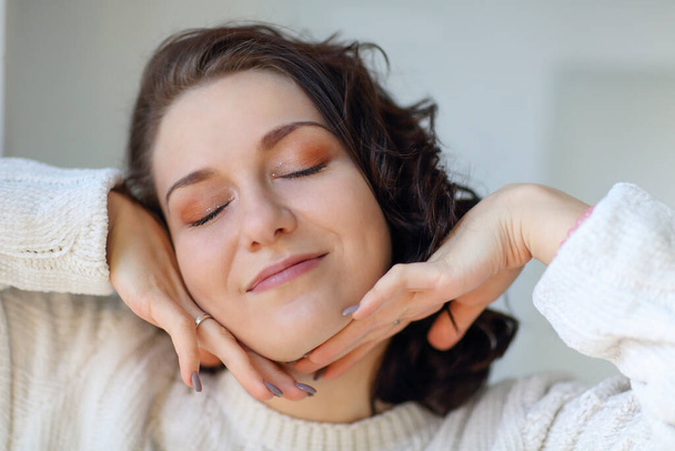 Close portrait of young happy pleased brunette girl with closed eyes touching clean fresh skin, putting hands on her face showing freshly done daily makeup, enjoying new day at home. Pleasure concept - 写真・画像
