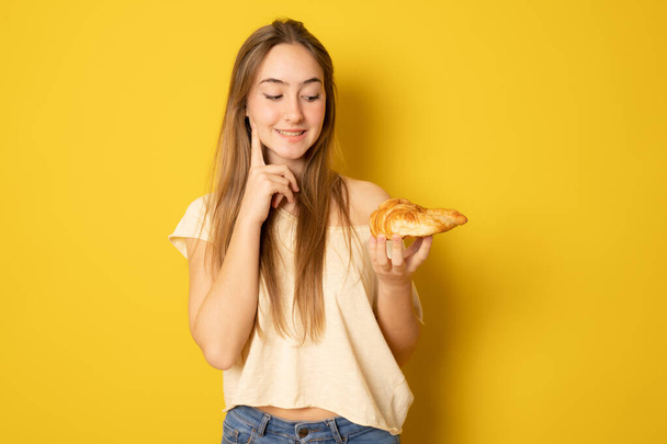 Young woman eating croissant, coffee break. Isolated over yellow background. Diet concept. - 写真・画像