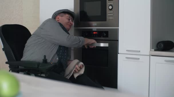 Cerrado de hombre caucásico en silla de ruedas con anteojos y gorra, abriendo la puerta del horno durante el día en su cocina moderna en casa - Imágenes, Vídeo
