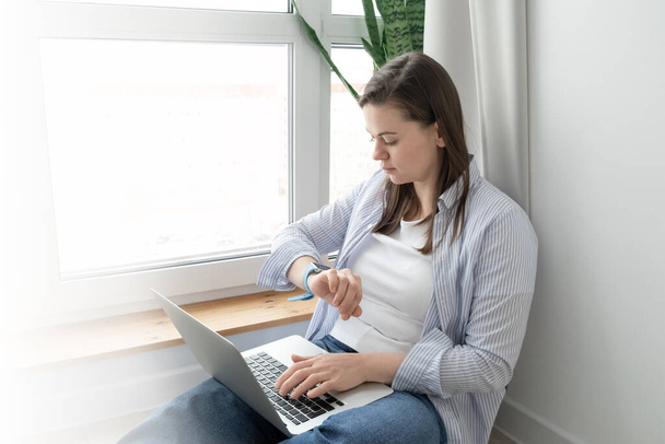 Mujer de mediana edad que trabaja desde casa. Chica usando el ordenador portátil en casa, sentado cerca de una gran ventana. Concepto de trabajo remoto - Foto, Imagen