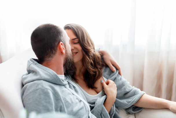 居心地の良い家のインテリアでソファに座っている間、若い美しいカップル抱擁キス。既婚男性と女性のロマンチックな関係。夫婦は家でお互いへの優しさの情熱を示す - 写真・画像
