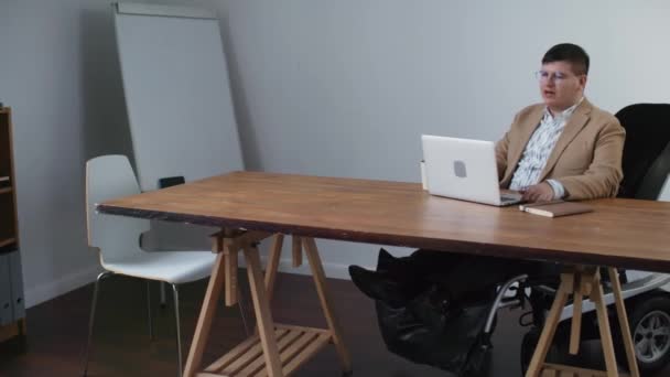 Wide shot of intelligent Caucasian businessman with disabilities dressed formally, sitting at desk in office at daytime, video calling via portable computer - Imágenes, Vídeo