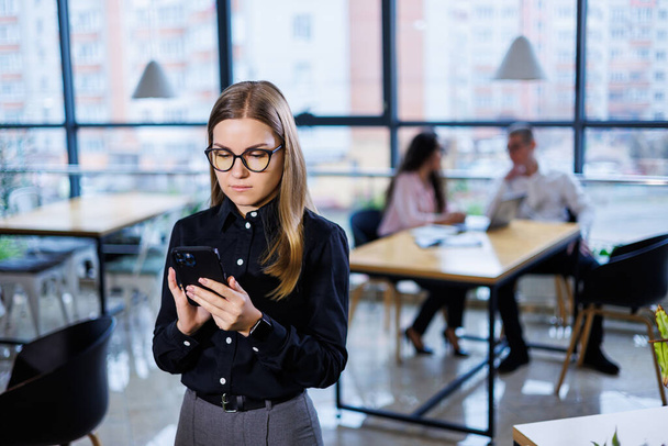 Γοητευτική επιχειρηματίας με γυαλιά καθημερινά, χρησιμοποιώντας smartphone και μιλώντας στο τηλέφωνο, σε μοντέρνο γραφείο. - Φωτογραφία, εικόνα