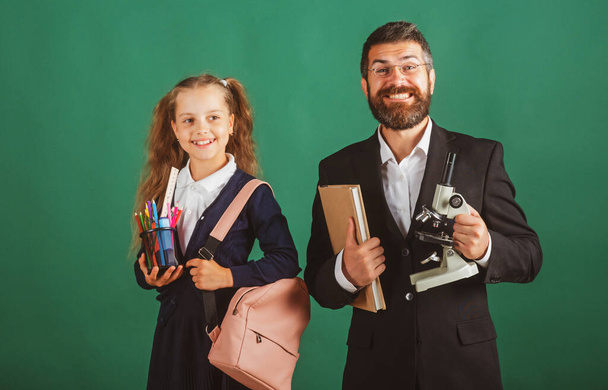 School girl with funny amazed teacher in school. Studio portrait of tutor and young school girl with backpack and textbook on blackboard green background. - Foto, Bild