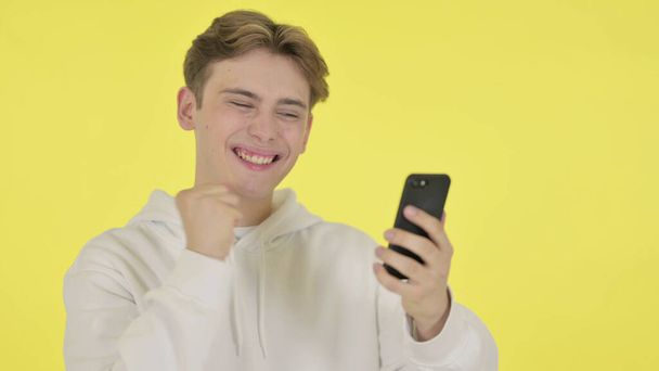 黄色の背景にスマートフォンで祝う若い男 - 写真・画像