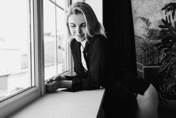 Portrait of a black and white young, pretty European girl emotional in a men's shirt in the bedroom near a large window, smiles, straightens hair. Morning sun, selective focus  - Foto, immagini