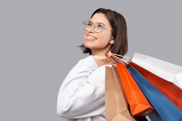 Shopping time. Close up photo of young trendy woman smiling and holding paper shopping bags over gray background - Foto, imagen