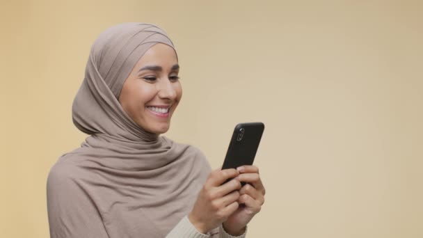 Διαδικτυακή επικοινωνία. Θετική νεαρή μεσανατολική κυρία με hijab γραπτών μηνυμάτων ή web surfing σε smartphone, ελεύθερο χώρο - Πλάνα, βίντεο