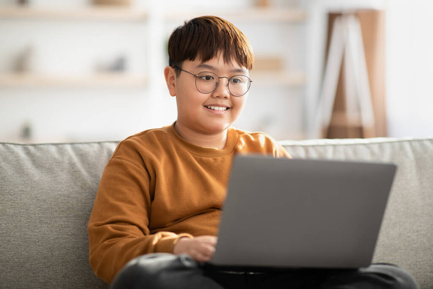 Alegre niño chino sentado en el sofá con el ordenador portátil en casa - Foto, Imagen