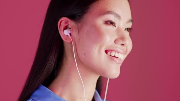 Χαρούμενη νεαρή ασιατική γυναίκα ακούγοντας μουσική σε ακουστικά πάνω από ροζ φόντο στούντιο - Πλάνα, βίντεο