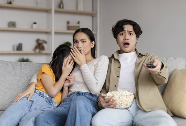 Frightened shocked asian millennial wife, husband and adolescent daughter watching scary movie and eating popcorn - Foto, afbeelding