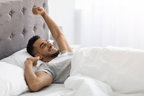 Τεμπέλικο πρωινό. Ευχαριστημένος μεσανατολίτης άντρας ξαπλωμένος στο κρεβάτι μετά τον ωραίο ύπνο - Φωτογραφία, εικόνα