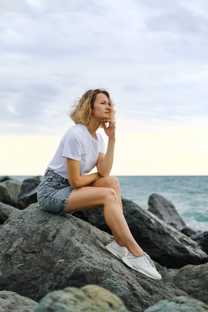 A young attractive Caucasian blonde woman of 35 years sits on the rocks of sea coast. Emerald-colored water, waves splashing on the stones. The woman meditates and relaxes. - Foto, Imagem