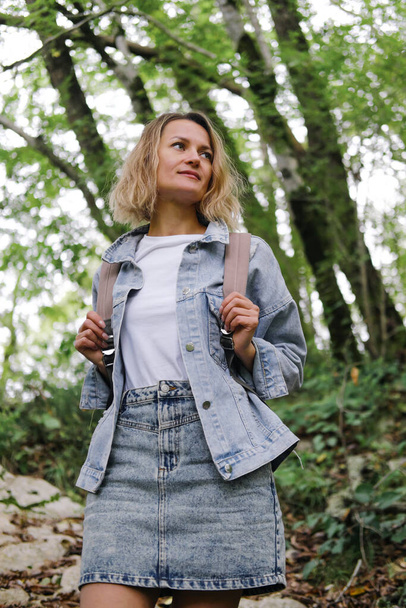 A young Caucasian woman walks along a forest mountain road. Casual blue denim clothes and backpack. Travel, healthy lifestyle and freedom. - 写真・画像