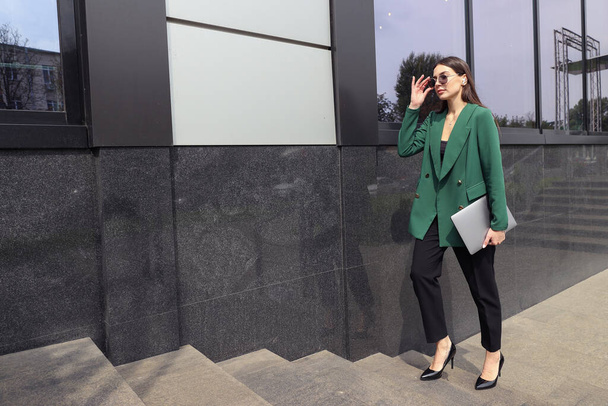 Una giovane donna in giacca e cravatta con un computer portatile sta camminando vicino a un edificio per uffici - Foto, immagini