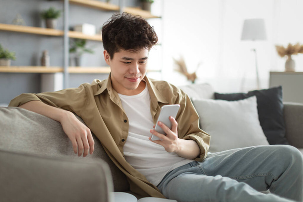 Smiling asian man using smartphone at home sitting on couch - Foto, immagini