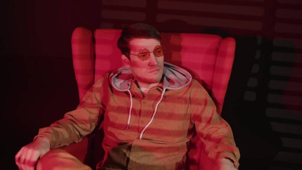 Προσεκτικός άνθρωπος κάθεται στην πολυθρόνα σε σκοτεινό δωμάτιο με κόκκινο φως που πέφτει και σκιές blinds. Σκεπτικός άντρας με γυαλιά κοιτάζει μακριά και σκέφτεται τη ζωή. - Φωτογραφία, εικόνα