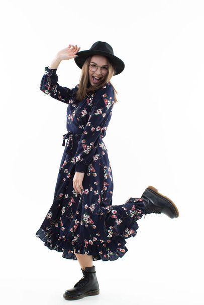 A brunette in a long floral dress with a hat poses for the camera in the studio. Emotional portrait - 写真・画像