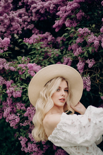 Young blonde girl in a straw hat and in a white wedding dress with an open top near the lilac bushes - Φωτογραφία, εικόνα