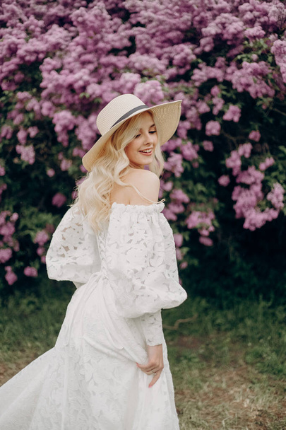 Young blonde girl in a straw hat and in a white wedding dress with an open top near the lilac bushes - Photo, Image