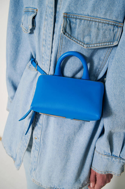 Woman in blue jeans shirt with small blue handbag on her body. Blue leather clutch with handle. Vertical photo closeup. - Photo, image