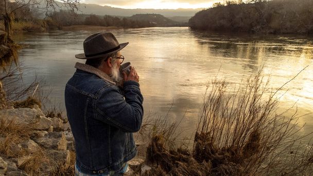 Hiker man enjoys beautiful natural scenery with a cup of hot drink. Man drinking coffee and admiring the beautiful river at sunrise. - Fotó, kép