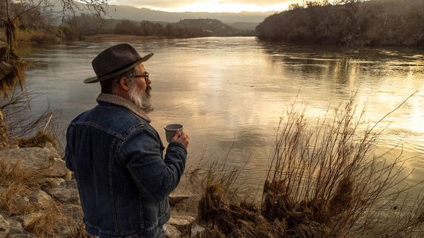 ハイカーの男は熱い飲み物のカップで美しい自然の風景を楽しんでいます。男はコーヒーを飲み、日の出に美しい川を賞賛. - 写真・画像