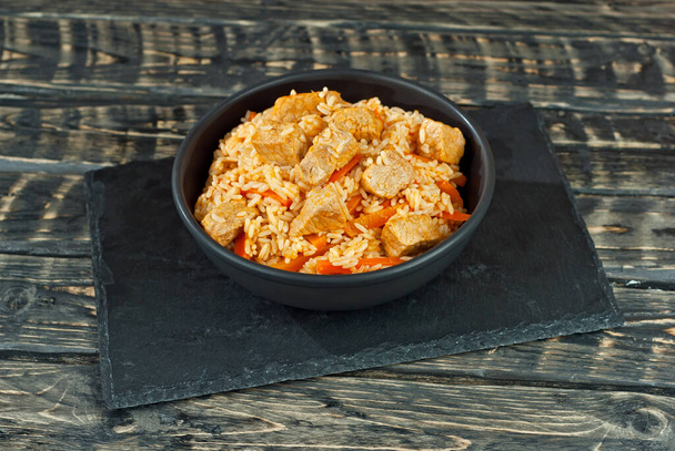 Pilaf with meat and carrots in a black bowl. Pilaf on a black background. Food on a shabby table. Bowl on an old wooden board. - Фото, изображение