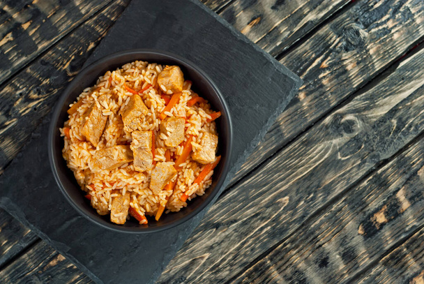 Pilaf with meat and carrots in a black bowl. Pilaf on a black background. Food on a shabby table. Bowl on an old wooden board. Copy space and free space for text near food. - Foto, Imagem