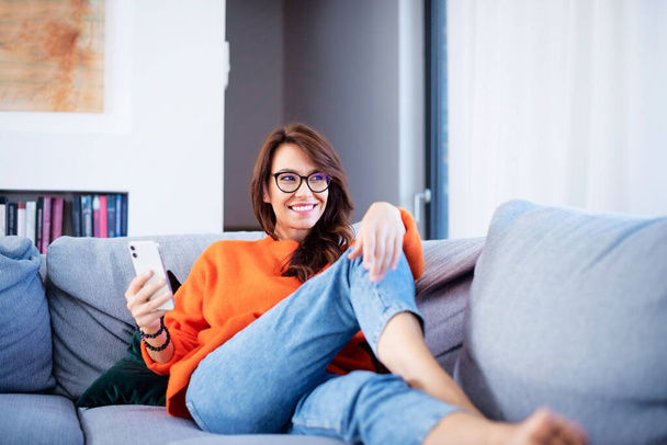 Shot of an attractive and happy woman using her smartphone and text messaging while relaxing on the couch at home.  - Photo, image