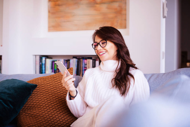 Shot of an attractive and happy woman using her smartphone and text messaging while relaxing on the couch at home.  - Photo, Image