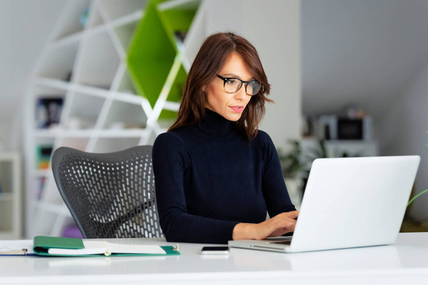 Confident middle aged businesswoman wearing turtleneck sweater while sitting at office desk behind her laptop and working on new project.  - Photo, Image