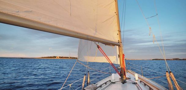 White sloop rigged yacht sailing at sunset. Clear sky after the storm. View from the deck to the bow, mast, sails. Transportation, travel, cruise, sport, recreation, leisure activity, racing, regatta - Fotó, kép