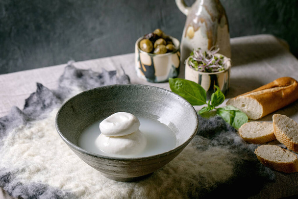 Traditional italian burrata knotted cheese in grey ceramic bowl on table. Bread, olives, green salad around. Ingredients for healthy mediterranean dinner. - Fotoğraf, Görsel