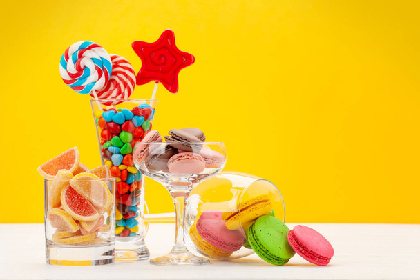 Diverse zoetigheden assortiment. Snoep, bonbon, chocolade en macarons in cocktailglazen op gele achtergrond. met kopieerruimte - Foto, afbeelding