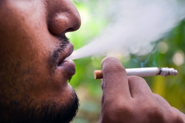 Молодой человек курит и задыхается. Сигарета зажжена и пепел на краю сигареты. Крупный план человека с сигаретой в руке и дымом изо рта. - Фото, изображение