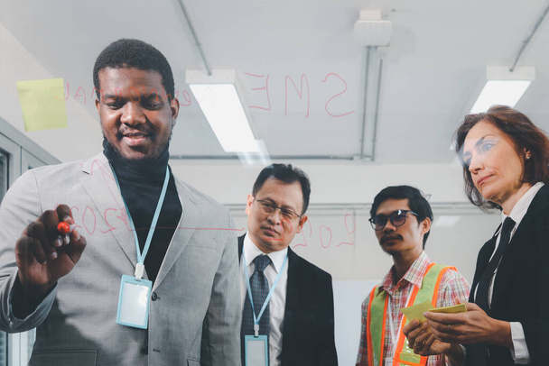 Young African businessman in suit with pen writing on glass board for creative brainstorming idea in office, critical thinking in project work, corporate colleague discussing for result, variety multi-ethnic team colleagues work together - Photo, image