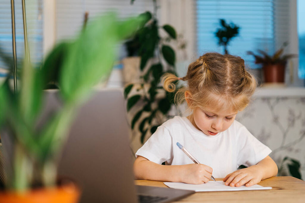 piccola ragazza bionda carina scrive, studia a casa davanti a un computer portatile, apprendimento online, torna a scuola - Foto, immagini
