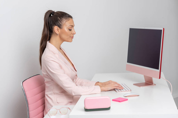 Egy nő ül egy munkaállomáson billentyűzet és asztali számítógép segítségével. Rózsaszín témájú. - Fotó, kép