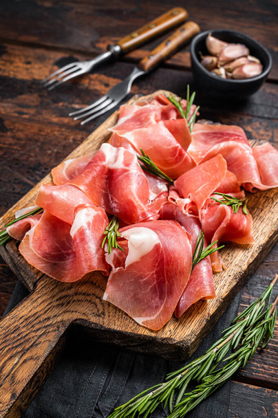 Slices of jamon serrano ham or prosciutto crudo parma on wooden board with rosemary. Wooden background. Top view - Photo, image